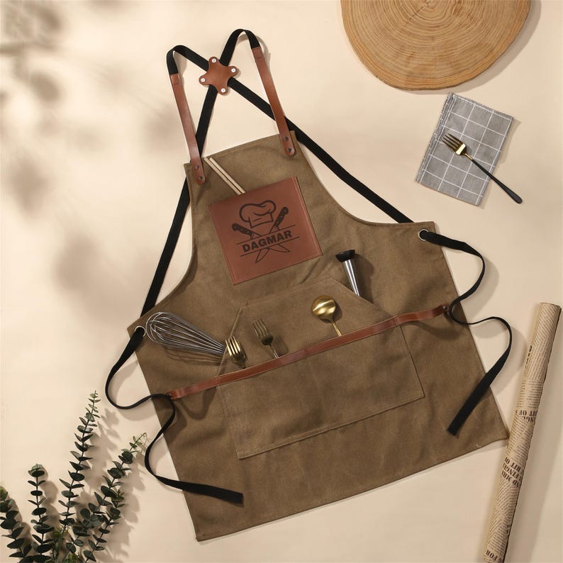 May include: Brown canvas apron with leather straps and pockets. Features a leather patch with the name "DAGMAR" and a chef's hat logo. Includes a whisk, forks, spoon, and other kitchen tools.