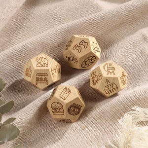 Pu&ograve; includere: Quattro dadi dodecaedrici in legno con icone incise. Ogni dado presenta diverse illustrazioni, tra cui un hamburger, uno stadio, controller di videogiochi e un barbecue. I dadi sono in legno di colore chiaro e poggiano su un tessuto dai toni neutri.