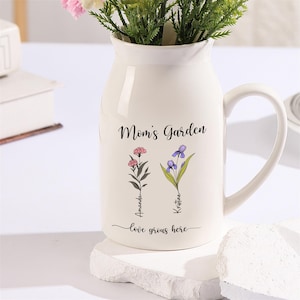 Może przedstawiać: Biały ceramiczny wazon z uchwytem, z napisem "Mom's Garden" i dwoma stylizowanymi ilustracjami kwiatowymi, pod którymi znajdują się imiona "Amanda" i "Kristine". Pod kwiatami znajduje się napis "love grows here".