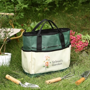 May include: A green and cream-colored garden tote bag with black handles and trim, featuring a floral design and the text "Selena's Garden." Garden tools, including a trowel and hand rake, are visible in the grass.