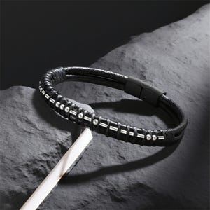May include: Black leather bracelet with silver-colored beads and accents. The bracelet features a magnetic clasp and a braided design. The accessory is displayed on a dark, textured surface.