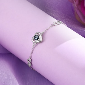May include: A silver bracelet with a heart-shaped charm that projects an image when held up to the light. The charm has a black pearl in the center.