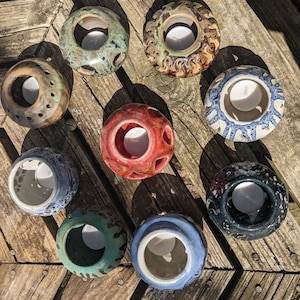 May include: Nine ceramic candle holders in various colors and patterns. The holders are round and have a hole in the center for a tea light candle. The holders are arranged in a circle on a wooden surface.