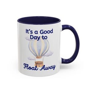 May include: A white ceramic mug with a dark blue interior and handle. The front features a watercolor illustration of a light blue and cream hot air balloon with clouds and the text "It's a Good Day to Float Away" in dark blue lettering.
