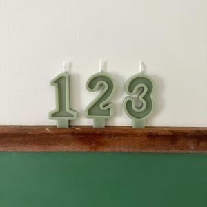 May include: Three sage green candles shaped like the numbers 1, 2, and 3. The candles are sitting on a wooden surface in front of a green chalkboard.