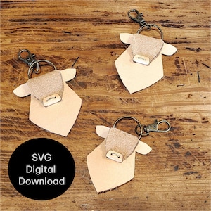 May include: Three cow-shaped leather keyrings with metal fixings are arranged on a wooden surface. Each keyring has a light tan body, a textured beige head, and a metal clasp. A black circle with white text reads "SVG Digital Download."