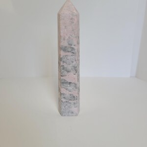 May include: A rectangular crystal obelisk with a pointed top. The stone has a mix of pink and grey tones, creating a marbled effect. The obelisk is a decorative item, likely used for display or spiritual purposes.