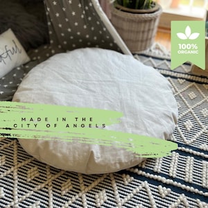 Könnte beinhalten: Ein weißes rundes Kissen mit dem Text "MADE IN THE CITY OF ANGELS" darauf. Das Kissen liegt auf einem schwarz-weiß gemusterten Teppich.