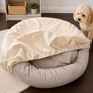 May include: A round dog bed with a removable, light beige cover. The bed's base is a muted gray color. A small, tan-colored dog stands nearby, looking at the bed. The bed is on a light-colored rug.