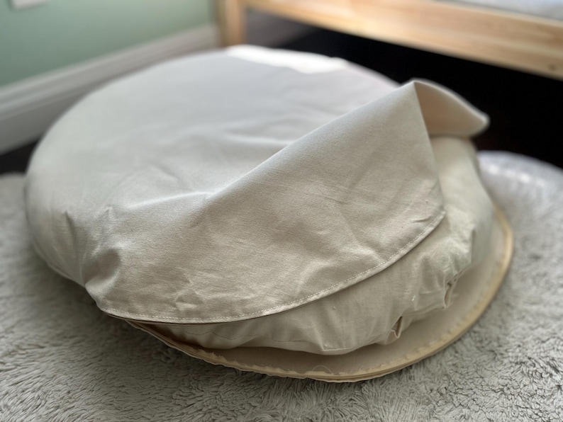 Natural cotton canvas dog bed cover partially lifted to reveal the inner cushion, displayed on a soft textured surface in a softly lit room