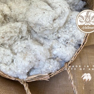 Puede incluir: Una cesta de mimbre llena de fibras de algodón blancas. La cesta tiene un asa de cuerda marrón y el texto "MADE IN THE CITY OF ANGELS" es visible en la esquina inferior derecha de la imagen. Un pequeño círculo marrón con el texto "Natural" es visible en la esquina superior derecha de la imagen.