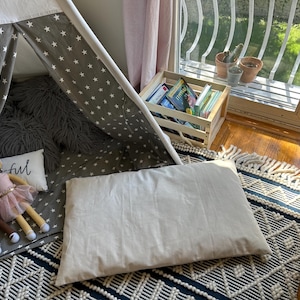 Puede incluir: Una tienda de campaña tipi gris y blanca con una alfombra gris esponjosa en el interior. Hay dos muñecas dentro de la tienda y una almohada blanca con la palabra "Joyful" en ella. Una almohada blanca grande está en el suelo fuera de la tienda. Una caja de madera con libros está en el fondo.