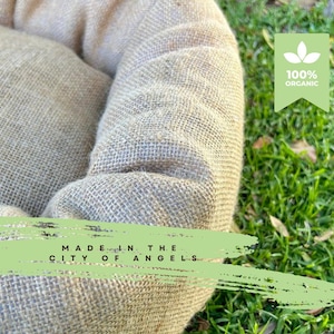 Puede incluir: Una cama redonda para mascotas hecha de yute natural, de color beige y tejido. La cama tiene un borde elevado y está colocada sobre una superficie de hierba verde. El texto "MADE IN THE CITY OF ANGELS" está escrito en la cama. Una etiqueta verde con el texto "100% ORGÁNICO" es visible en el fondo.