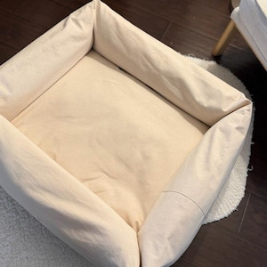 May include: A beige, rectangular pet bed with raised sides. The bed is made of a soft, light-colored fabric and sits on a white, fluffy rug. The bed is empty and ready for a pet to use.