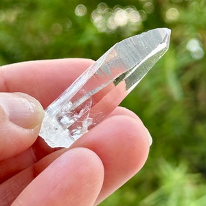 May include: A clear quartz crystal point with a double termination. The crystal is held in a hand against a blurred green background.