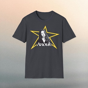May include: Dark gray t-shirt featuring a yellow star design with a portrait of a woman and the name "Anouk" in white script. The t-shirt has short sleeves and a crew neck.