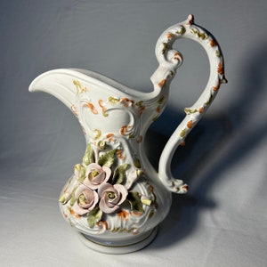 Peut inclure: Un pichet en porcelaine blanche avec un motif floral. Le pichet a une anse et un bec verseur. Le motif floral présente des roses roses et des feuilles vertes.