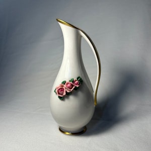 May include: White ceramic vase with a gold rim and handle. The vase features a floral design with pink roses and green leaves.