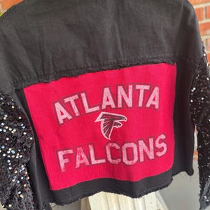 May include: Black denim jacket with a red patch on the back that says "Atlanta Falcons". The jacket has sequined sleeves.