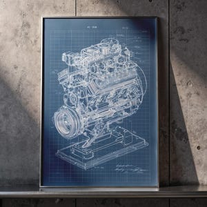 May include: A framed blueprint-style illustration of an engine, rendered in white lines against a blue grid background. The detailed technical drawing is encased in a silver frame, showcasing the engine's components. The print is displayed on a dark surface against a concrete wall.