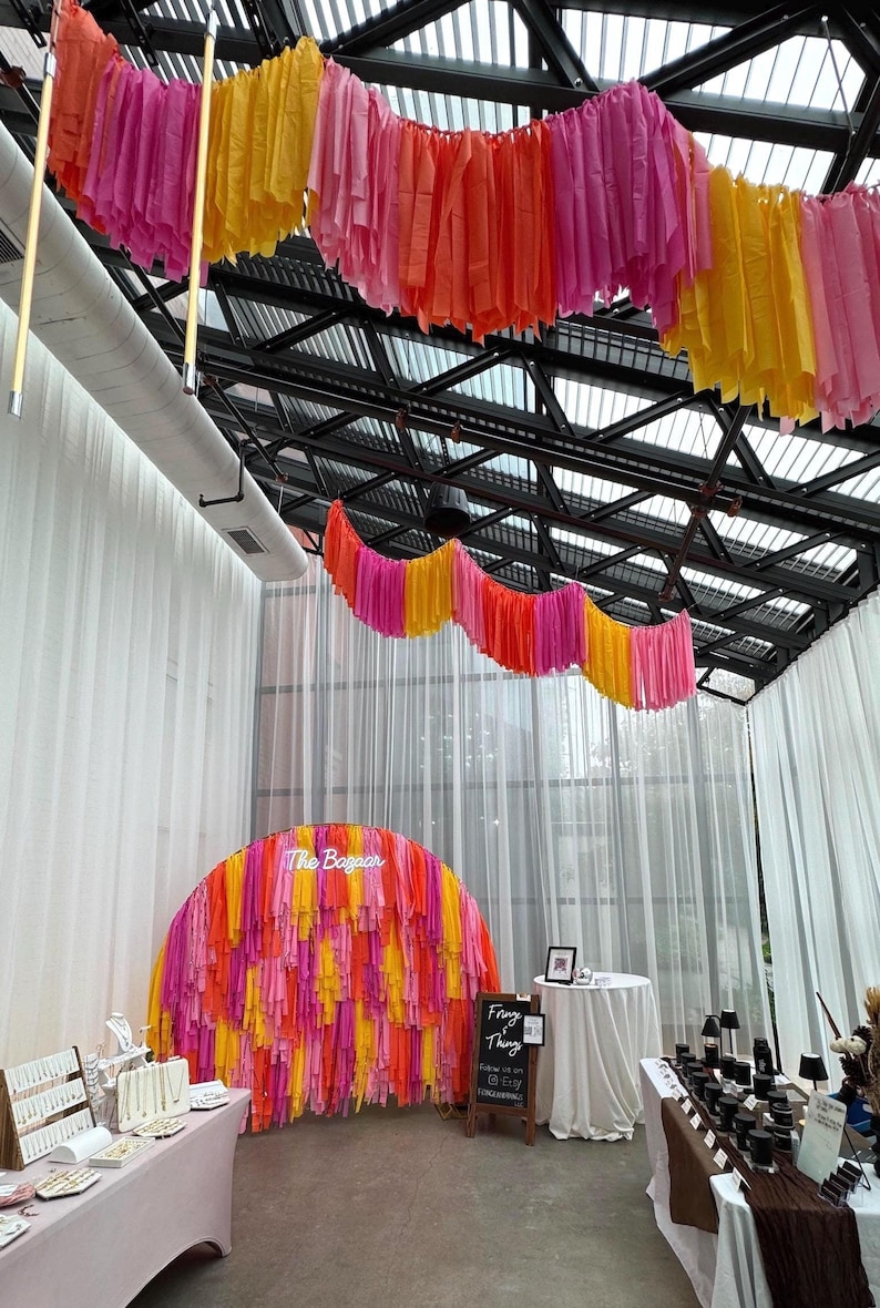 May include: A market stall with a pink, yellow, and orange fringe backdrop. The stall is decorated with a neon sign that says "The Bazaar". There is a sign that says "Fringe Thing" and a table with jewelry on display.
