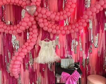 May include: A pink and silver balloon arch with a white dress hanging in front of it. The balloon arch is made of pink balloons and is shaped like a heart. The dress is a white tulle dress with a pink bow on the front. The balloon arch is hanging on a wall that is covered in pink and silver streamers.