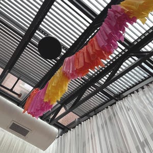 May include: A black metal ceiling with a white and gray striped awning. A colorful paper garland hangs from the ceiling, with pink, orange, and yellow paper streamers. White sheer curtains hang on the sides of the room.