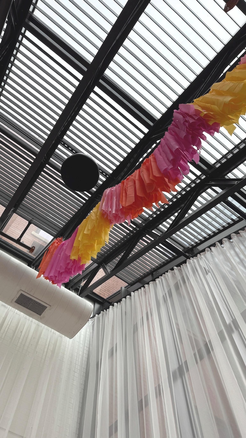 May include: A black metal ceiling with a white and gray striped awning. A colorful paper garland hangs from the ceiling, with pink, orange, and yellow paper streamers. White sheer curtains hang on the sides of the room.
