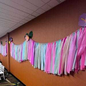 Pode incluir: Uma guirlanda de papel crepom colorido com serpentinas rosa, azul e verde pendura em uma parede marrom. A guirlanda &eacute; decorada com flores de papel, incluindo um grande hibisco roxo.