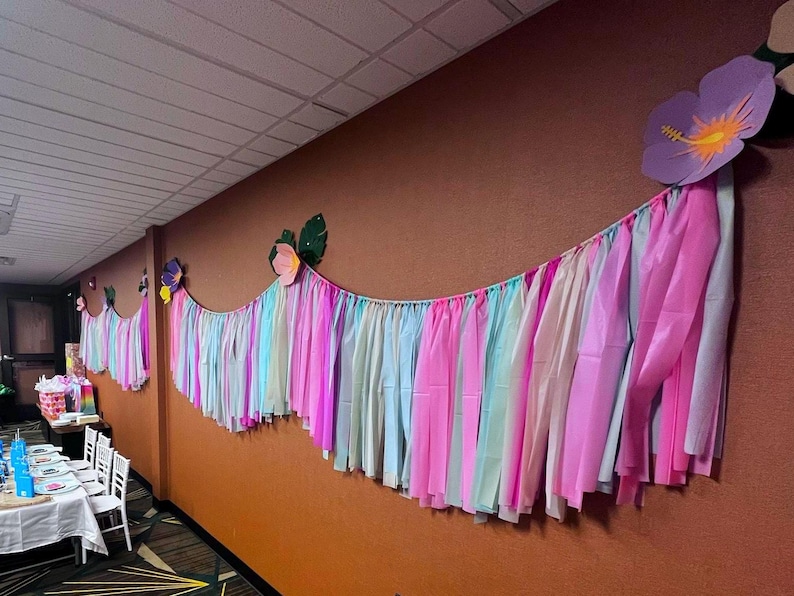 Pode incluir: Uma guirlanda de papel crepom colorido com serpentinas rosa, azul e verde pendura em uma parede marrom. A guirlanda &eacute; decorada com flores de papel, incluindo um grande hibisco roxo.