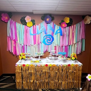 Pode incluir: Uma mesa decorada para uma festa com uma placa azul e branca "Liana", um desenho de espiral azul e uma saia de grama marrom. A mesa &eacute; decorada com serpentinas de papel coloridas, flores e sobremesas.