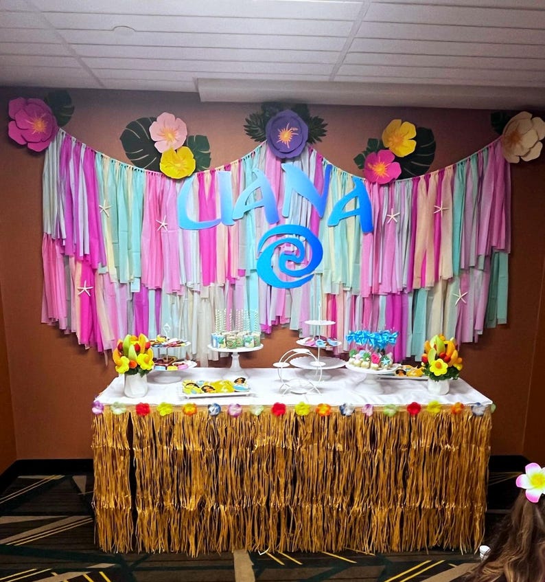 Pode incluir: Uma mesa decorada para uma festa com uma placa azul e branca "Liana", um desenho de espiral azul e uma saia de grama marrom. A mesa &eacute; decorada com serpentinas de papel coloridas, flores e sobremesas.