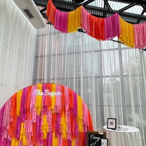 May include: A neon sign that says "The Bazaar" is in front of a backdrop made of pink, orange, and yellow fringe. A chalkboard sign says "Fringe & Things" and includes the text "Follow us on @Etsy FRINGEANDTHINGS LLC".