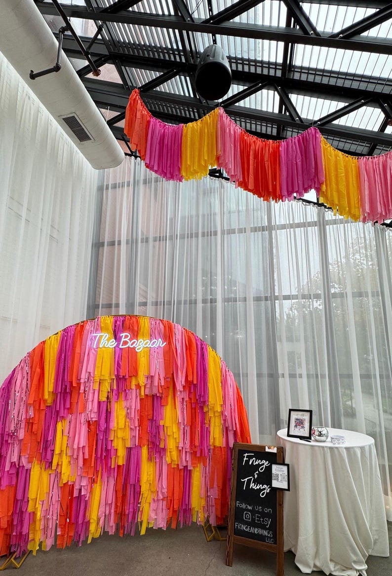 May include: A neon sign that says "The Bazaar" is in front of a backdrop made of pink, orange, and yellow fringe. A chalkboard sign says "Fringe & Things" and includes the text "Follow us on @Etsy FRINGEANDTHINGS LLC".