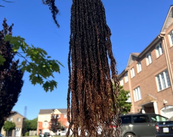 Op de afbeelding: Een lange set zwarte en bruine ombre dreadlocks van echt haar. De dreadlocks worden in een hand gehouden en zijn ongeveer 60 cm lang.