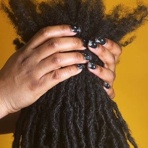 May include: A person's hand holding a thick, dark brown dreadlock hairstyle with black nail polish and white glitter stars.