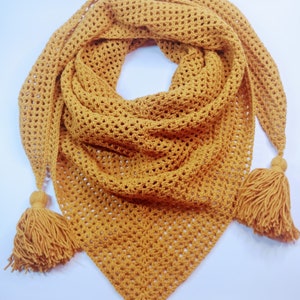May include: Mustard yellow crochet shawl with a triangular shape. The shawl features a delicate openwork pattern and is finished with tassels at each point. The handmade scarf is a cozy accessory.