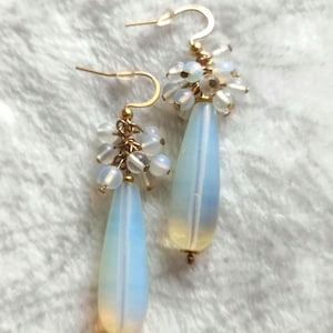 Könnte beinhalten: Ein Paar Ohrhänger. Jeder Ohrring hat einen goldfarbenen Haken, eine Ansammlung kleiner, runder, weißer Perlen und eine tropfenförmige, opaleszierende Perle mit einem Farbverlauf von Blau nach Gelb.