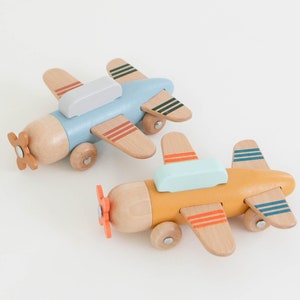 K&ouml;nnte beinhalten: Zwei Holzspielzeugflugzeuge. Eines ist hellblau mit einem grauen Oberteil und das andere ist gelb mit einem hellgr&uuml;nen Oberteil. Beide haben Holzpropeller und R&auml;der. Die Fl&uuml;gel haben farbige Streifen.