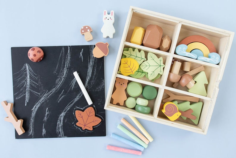 Pode incluir: Uma caixa de madeira cheia de brinquedos de madeira coloridos, incluindo um urso, folhas, cogumelos e um arco-&iacute;ris. A caixa est&aacute; aberta e os brinquedos s&atilde;o vis&iacute;veis. H&aacute; tamb&eacute;m um quadro-negro com um giz e alguns brinquedos de madeira nele.