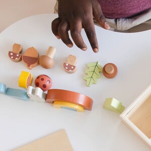 Pode incluir: Um conjunto de brinquedos de madeira com v&aacute;rias formas e cores, incluindo cogumelos, bolotas, folhas e um arco-&iacute;ris. O conjunto est&aacute; disposto sobre uma superf&iacute;cie branca com uma caixa de madeira no fundo.