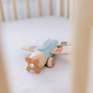 K&ouml;nnte beinhalten: Ein Holzspielzeugflugzeug mit hellblauem Rumpf, Fl&uuml;geln aus Naturholz und einem orangefarbenen Propeller. Die Fl&uuml;gel haben rote und schwarze Streifen. Das Spielzeug liegt auf einer wei&szlig;en Oberfl&auml;che, m&ouml;glicherweise einem Kinderbett.