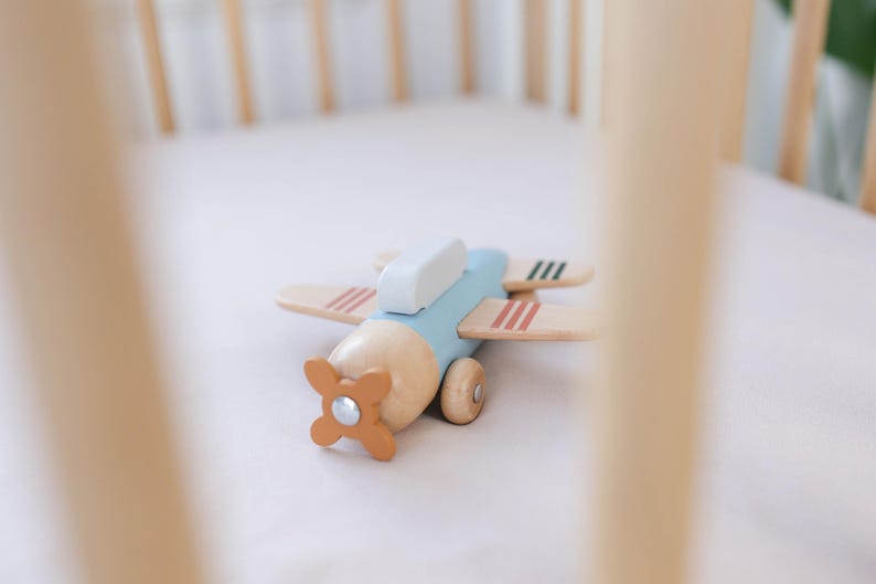 K&ouml;nnte beinhalten: Ein Holzspielzeugflugzeug mit hellblauem Rumpf, Fl&uuml;geln aus Naturholz und einem orangefarbenen Propeller. Die Fl&uuml;gel haben rote und schwarze Streifen. Das Spielzeug liegt auf einer wei&szlig;en Oberfl&auml;che, m&ouml;glicherweise einem Kinderbett.