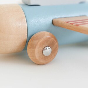 K&ouml;nnte beinhalten: Nahaufnahme eines Holzspielzeugflugzeugs. Das Flugzeug hat einen hellblauen K&ouml;rper, Akzente aus Naturholz und ein Holzrad. Der Fl&uuml;gel hat rote Streifen. Das Spielzeug ist f&uuml;r Kinder konzipiert.
