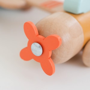 K&ouml;nnte beinhalten: Nahaufnahme eines Holzspielzeugflugzeugs. Der orangefarbene Propeller ist im Fokus, befestigt mit einem silbernen Metallteil. Der Rumpf des Flugzeugs ist hellbraun und gelb, mit einem hellblauen Akzentteil oben. Ein klassisches Spielzeug.