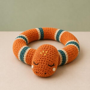 Peut inclure: Un jouet serpent en crochet de forme circulaire avec un corps orange brûlé et des rayures bleu sarcelle et crème. La tête du serpent présente des yeux fermés brodés, des taches blanches sur les joues et des détails jaunes sur le dessus.