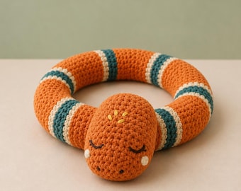 May include: A circular crochet snake toy featuring a burnt orange body with teal and cream striped accents. The snake head has embroidered closed eyes, small white cheek spots, and yellow stitch details on the top, crafted in yarn.
