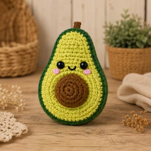 May include: A crocheted avocado plush toy featuring a dark green outer skin, a light yellow-green interior, and a central brown pit. The avocado has two black plastic eyes, a small stitched black smile, and two pink embroidered circular cheeks.