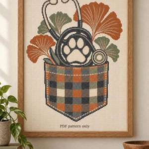 Può includere: Un motivo a punto croce raffigurante una tasca a quadri contenente uno stetoscopio con un disegno a forma di zampa, circondato da foglie di ginkgo arancioni e verdi. Il disegno è incorniciato in legno e include il testo PDF pattern only in basso.