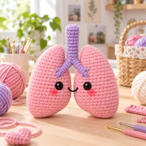 May include: A crochet plush toy shaped like a pair of human lungs, featuring light pink lobes and a lavender trachea. The lungs have black plastic safety eyes, a small embroidered black smile, and pink stitched cheeks, creating a cute appearance.